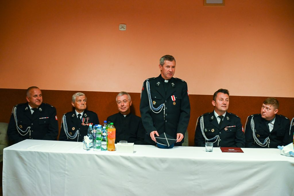 Sześciu mężczyzn w mundurach z odznaczeniami siedzi przy białym stole na tle pomarańczowej ściany, jeden stoi trzymając granatowy kapelusz.