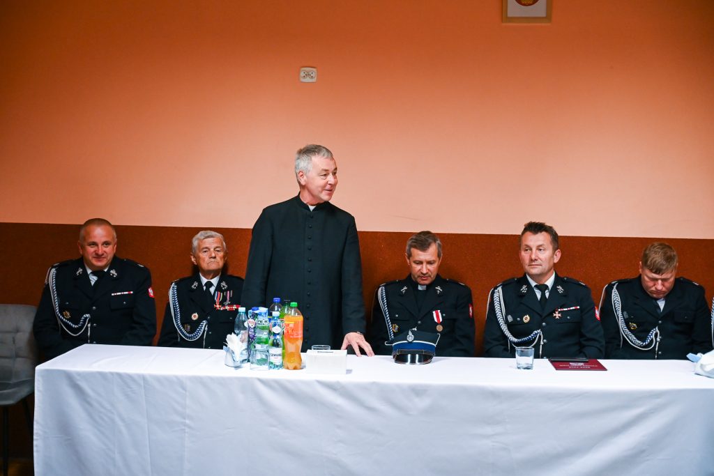 Pięciu mężczyzn w czarnych mundurach z odznaczeniami i jeden mężczyzna w czarnej sutannie przy stole z białym obrusem, na stole butelki i szklanki, tło pomarańczowo-brązowe.