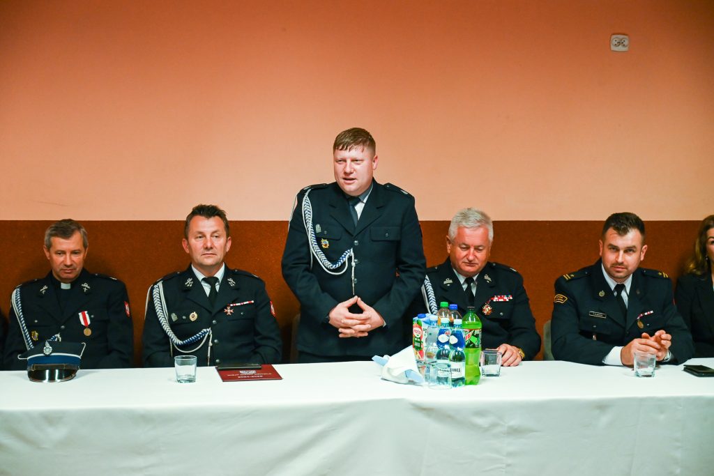 Piątka mężczyzn w mundurach, siedzący i stojący za stołem z białym obrusem, na stole butelki napojów i szklanki, pomarańczowo-brązowa ściana w tle.