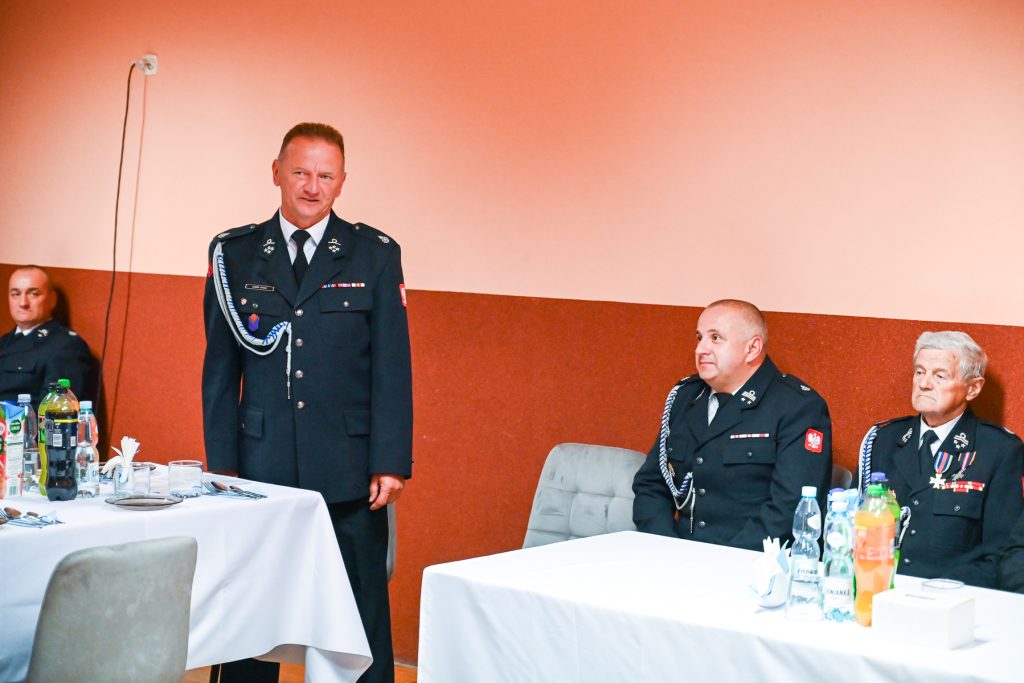 Trzech mężczyzn w mundurach strażackich siedzi i stoi przy stołach z białymi obrusami, na stołach butelki z napojami i szklanki, tło ściana w dwóch odcieniach czerwieni.