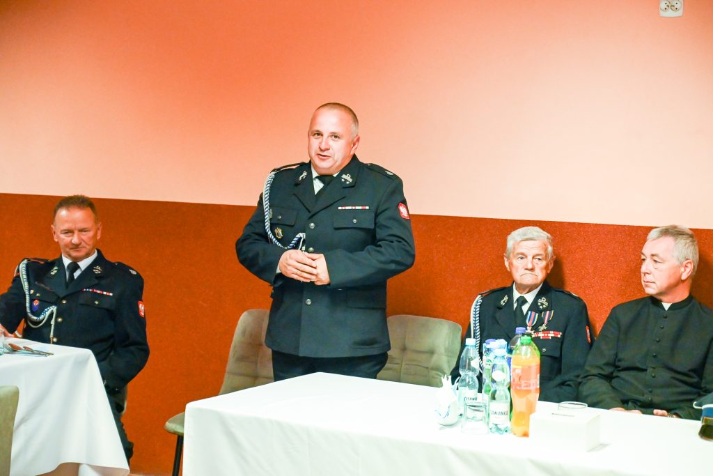 Cztery osoby w mundurach i czarnym stroju, trzy siedzą, jedna stoi, tło pomarańczowo-brązowe, na stole białe obrusy i butelki z napojami.
