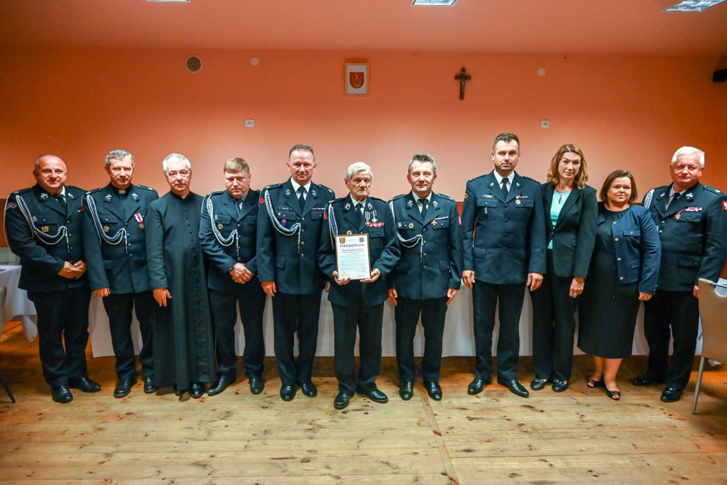Dwunastu ludzi w mundurach i garniturach stojących na drewnianej podłodze na tle pomarańczowej ściany z małym godłem i krzyżem.