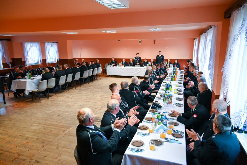 Mężczyźni w mundurach siedzą przy długich stołach z białymi obrusami, na stołach talerze, sztućce, napoje, pomarańczowe światełka, pomieszczenie z różowymi ścianami i białymi zasłonami.
