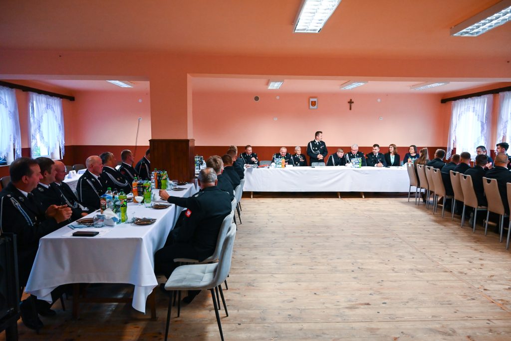Mężczyźni i kobiety w mundurach siedzą przy dwóch białych stołach w sali z różowymi ścianami i drewnianą podłogą, oświetlonej lampami sufitowymi, z zasłoniętymi firankami oknami.
