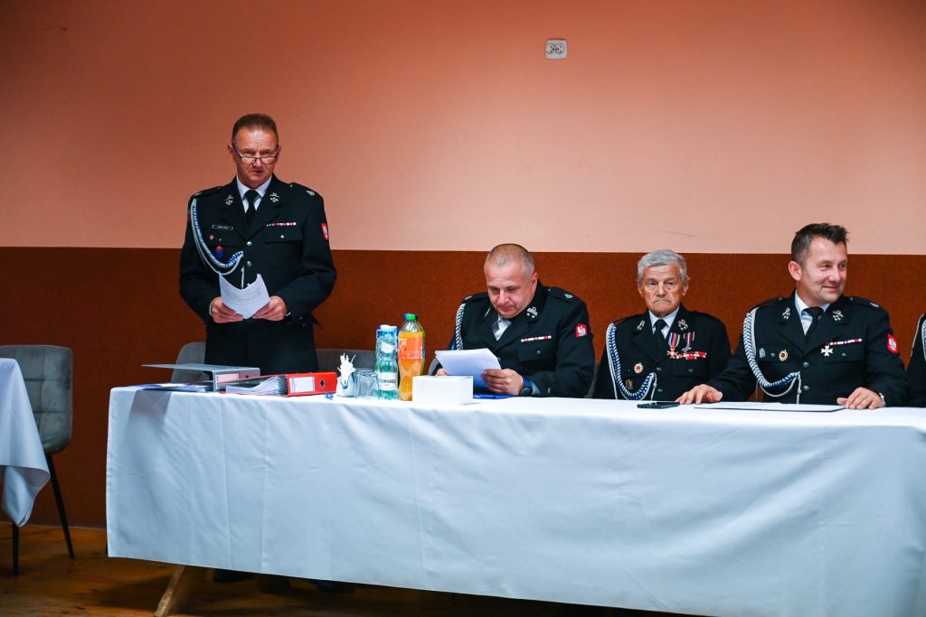 Czterech mężczyzn w czarnych mundurach z odznaczeniami siedzi i stoi przy białym stole na tle brązowo-pomarańczowej ściany, na stole butelki i dokumenty.