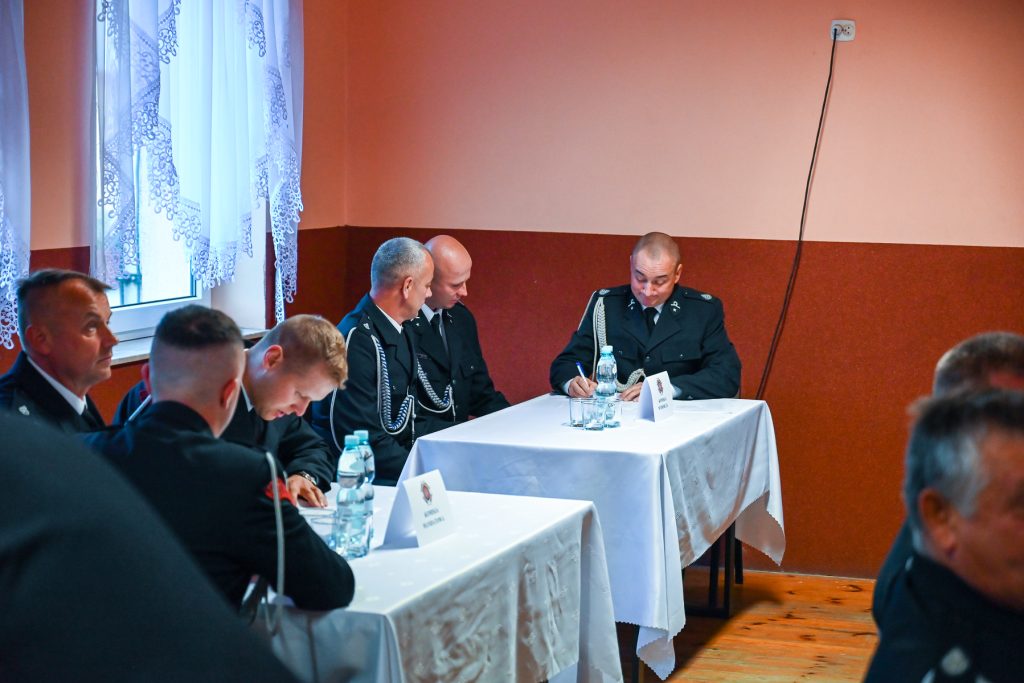 Mężczyźni w mundurach siedzą przy białych stołach w sali z pomarańczowymi ścianami, na stołach butelki wody i kartki z napisem.