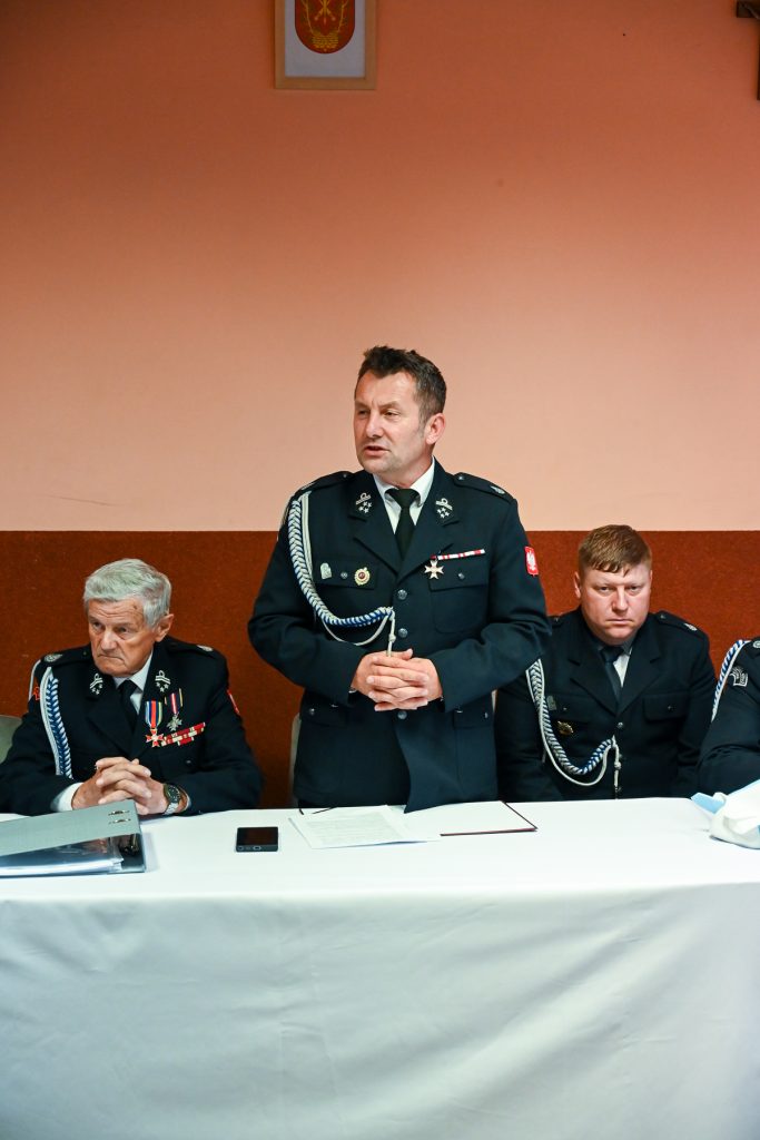 Trzech mężczyzn w mundurach z odznaczeniami siedzi i stoi przy stole z białym obrusem na tle pomarańczowo-brązowej ściany.